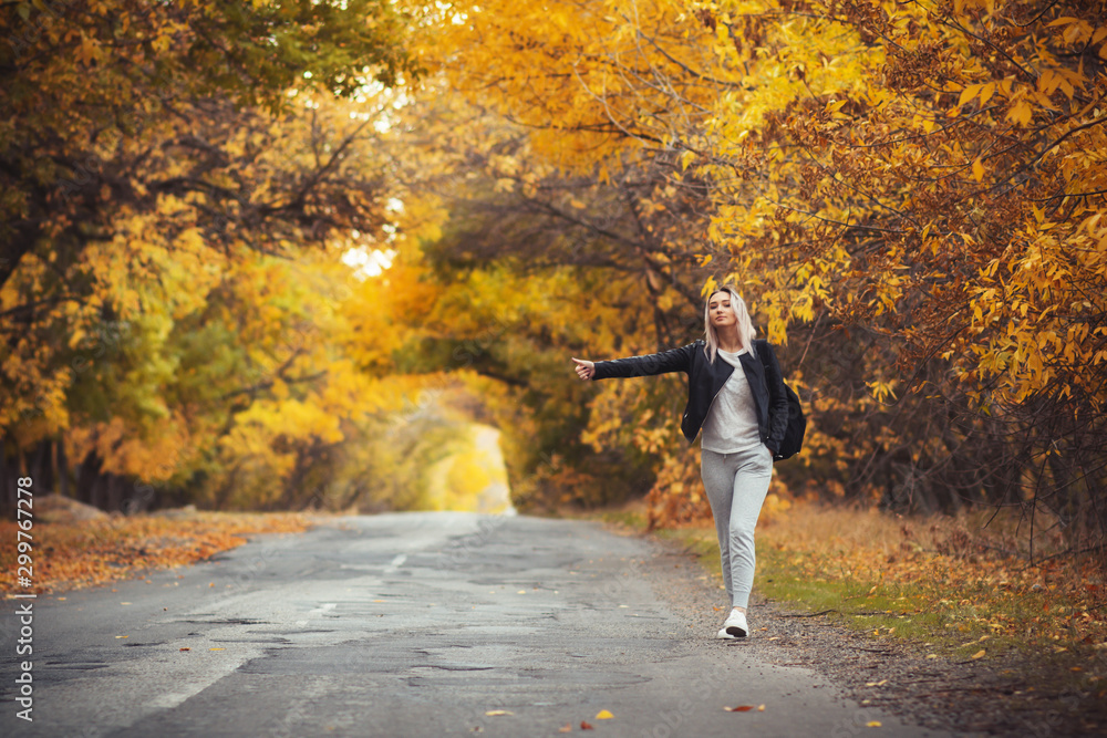 Obraz premium young woman stands and waits on an asphalt road in cold autumn weather, a girl tries to get from a suburban highway by a hitchhiker showing a hand gesture