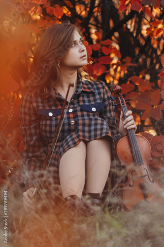 beautiful young woman sitting on the ground with a violin on a background of red foliage, romantic girl in dress inspired by nature for creative activity, concept of hobby and music