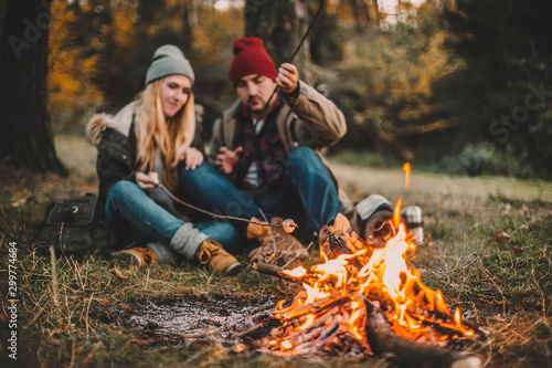Traveler couple camping and roasting marshmallows over the fire in the forest after a hard day. Concept of trekking, adventure and seasonal vacation.