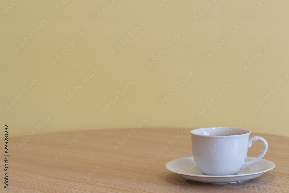 Obraz premium White coffee or tea cup with not soft stream smoke put on wooden table in a living room and blurry dark yellow cement wall with window lighting background. Selective focus and copy space.