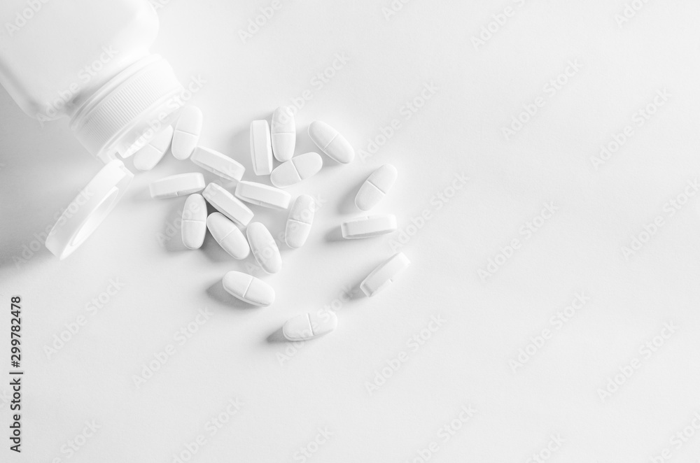 A white plastic container with white tablets lies on a white background next to white tablets A white plastic container with white tablets lies on a white background next to white tablets
