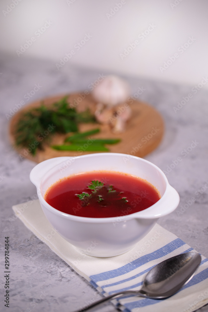 A plate with beetroot borsch on the table, next to the board is parsley, dill, green onions and garlic. Tasty and hearty lunch. A plate with beetroot borsch on the table, next to the board is parsley, dill, green onions and garlic. Tasty and hearty lunch.
