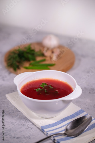 A plate with beetroot borsch on the table, next to the board is parsley, dill, green onions and garlic. Tasty and hearty lunch.
