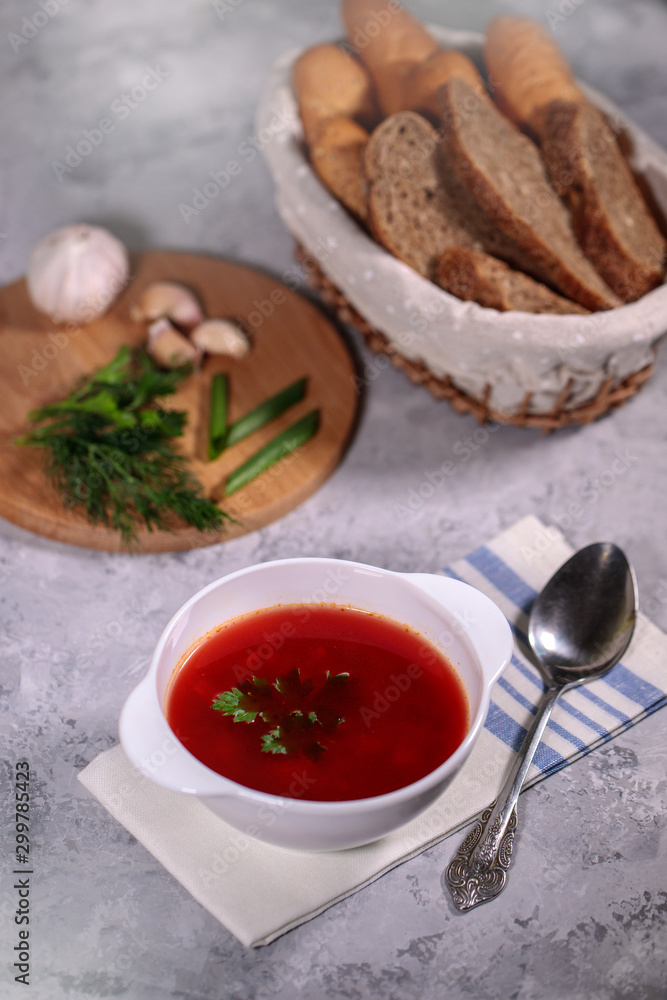 A plate with beetroot borsch on the table, next to the board is parsley, dill, green onions and garlic and a basket with bread. Tasty and hearty lunch. A plate with beetroot borsch on the table, next to the board is parsley, dill, green onions and garlic and a basket with bread. Tasty and hearty lunch.