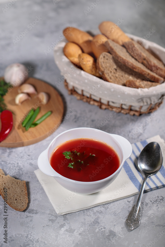 A plate with beetroot borsch on the table, next to the board is parsley, dill, green onions and garlic and a basket with bread. Tasty and hearty lunch. A plate with beetroot borsch on the table, next to the board is parsley, dill, green onions and garlic and a basket with bread. Tasty and hearty lunch.