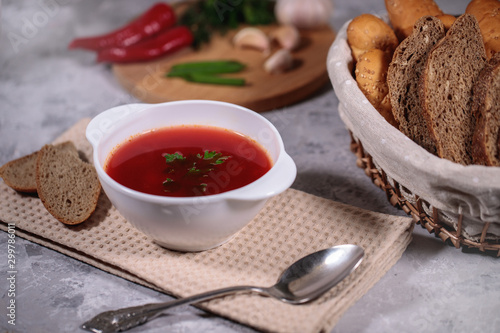 Tasty and hearty dinner. A plate with borsch on the table, next to the board is parsley, dill, green onions, garlic, chili pepper and a basket with bread.