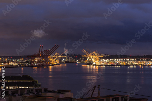 Purple water and sky surrounding large shipping cranes with lights as the sun sets