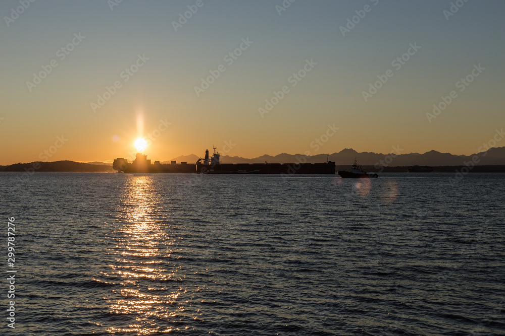 Sun setting right on top of a large container ship resting in the sound with a tug boat
