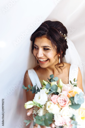 Young bride in white dress with a bouquet of flowers posing under curtain. Beautiful woman with professional make up and hair style. Morning of the bride. Wedding day. Fashion bride.