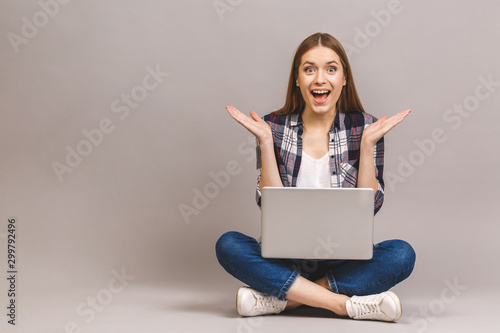 Wow, great news! Happy young amazed woman sitting on the floor with crossed legs and using laptop isolated on gray background. © denis_vermenko