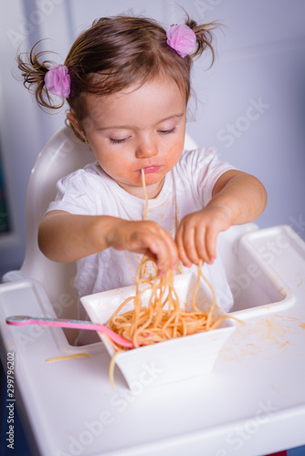 Cute little 1 year old girl  baby  child  sitting in a high-powered chair at home  eats noodles hands  photo in real life interior  self-feeding concept