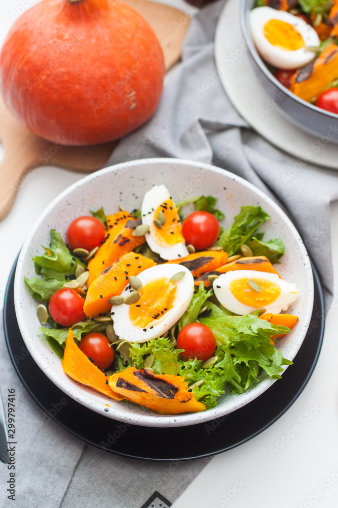 Lunch bowl with cherry tomatoes, roasted pumpkin, green salad, seeds and boiled eggs. Grey plate on white background. 