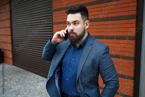 Stylish handsome brunette man with beard, wearing classic formal blue suit jacket, talking on a smartphone outdoors on the city street near garage roller door. Businessman using mobile phone.