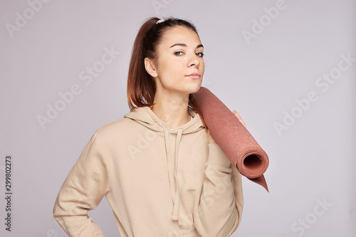 Headshot of serious thoughtful female teenager dressed in casual sweatshirt, holds mat, goes in for sport on gym, has active lifestyle, poses over grey blank wall indoor. Youth and recreation concept.