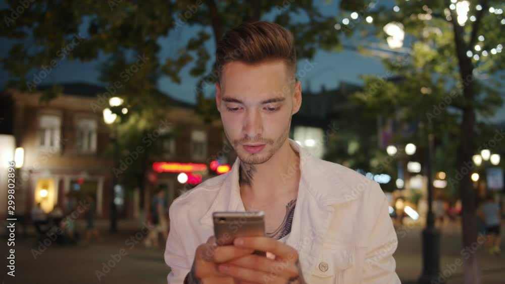 Slow motion of attractive student using smartphone touching screen outdoors standing in city street at night . Devices, people and modern youth concept. Slow motion of attractive student using smartphone touching screen outdoors standing in city street at night . Devices, people and modern youth concept.