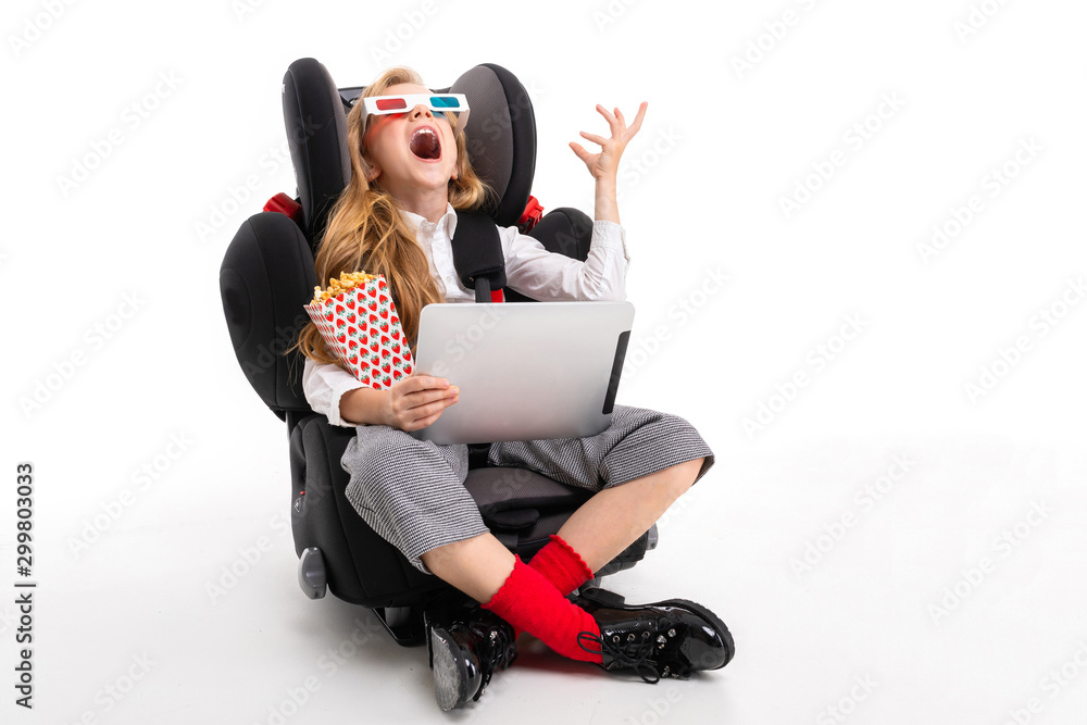 A little girl with makeup and long blonde hair sitting in a car baby chair  with tablet, earphones, pop-corn, 3D glasses and watch interesting comedy  film of cartoon Stock Photo | Adobe