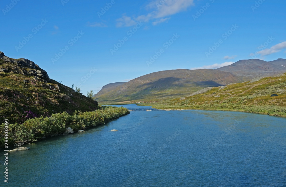 Lapland nature landscape with blue glacial lake Allesjok near Alesjaure, birch tree forest, snow capped mountains and sami village with red houses. Kungsleden hiking trail. Summer sunny day