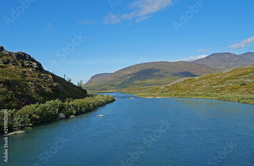 Lapland nature landscape with blue glacial lake Allesjok near Alesjaure, birch tree forest, snow capped mountains and sami village with red houses. Kungsleden hiking trail. Summer sunny day