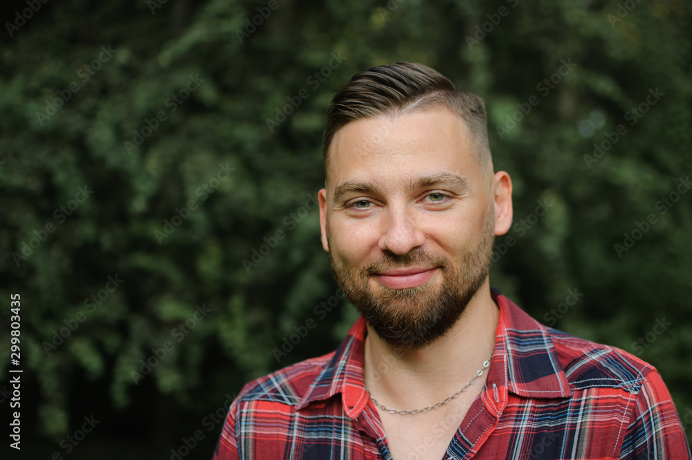 Close up portrait of young bearded man in red shirt looking at the camera with smile in the park on trees background Close up portrait of young bearded man in red shirt looking at the camera with smile in the park on trees background