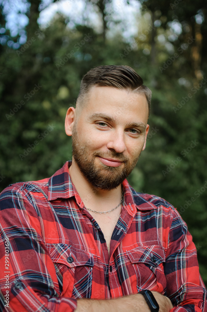 Close up portrait of young bearded man in red shirt looking at the camera with smile in the park on trees background Close up portrait of young bearded man in red shirt looking at the camera with smile in the park on trees background