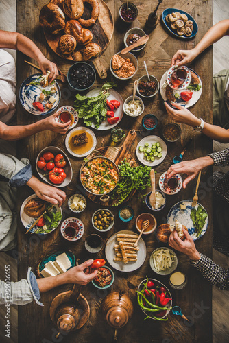 Turkish breakfast. Flat-lay of pastries, vegetables, greens, cheeses, fried eggs, jams in oriental tableware, tea in tulip glasses and peoples hands over rustic wooden background, top view