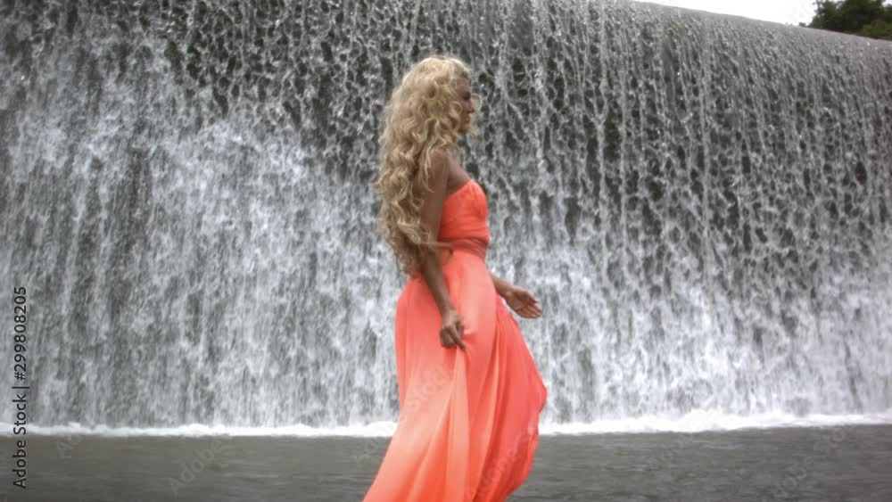 Young beautiful woman in orange dress walking and smiling on background of big waterfall. Girl turning head and looking in camera. Young beautiful woman in orange dress walking and smiling on background of big waterfall. Girl turning head and looking in camera.