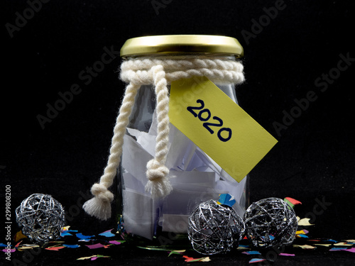 Glass jar full of paper shreds and tag reading 2020 surrounded by colorful confetti and silver ornaments isolated on black background