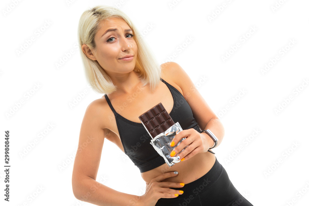 A young sports girl with blonde hair in a black sports top and black leggings holds chocolate and think A young sports girl with blonde hair in a black sports top and black leggings holds chocolate and think