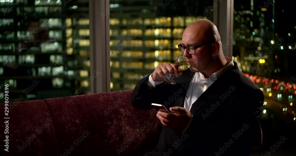 Portrait of a business man, he sits in the office on the couch at the panoramic window, behind which you can see the lights of the city, he holds a glass of wine in his hand and looks at the phone. Portrait of a business man, he sits in the office on the couch at the panoramic window, behind which you can see the lights of the city, he holds a glass of wine in his hand and looks at the phone.