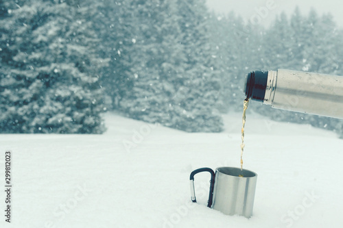 Tea from thermos pouring into a travel mug standing on the snow. Outdoors winter background with snowy spruce