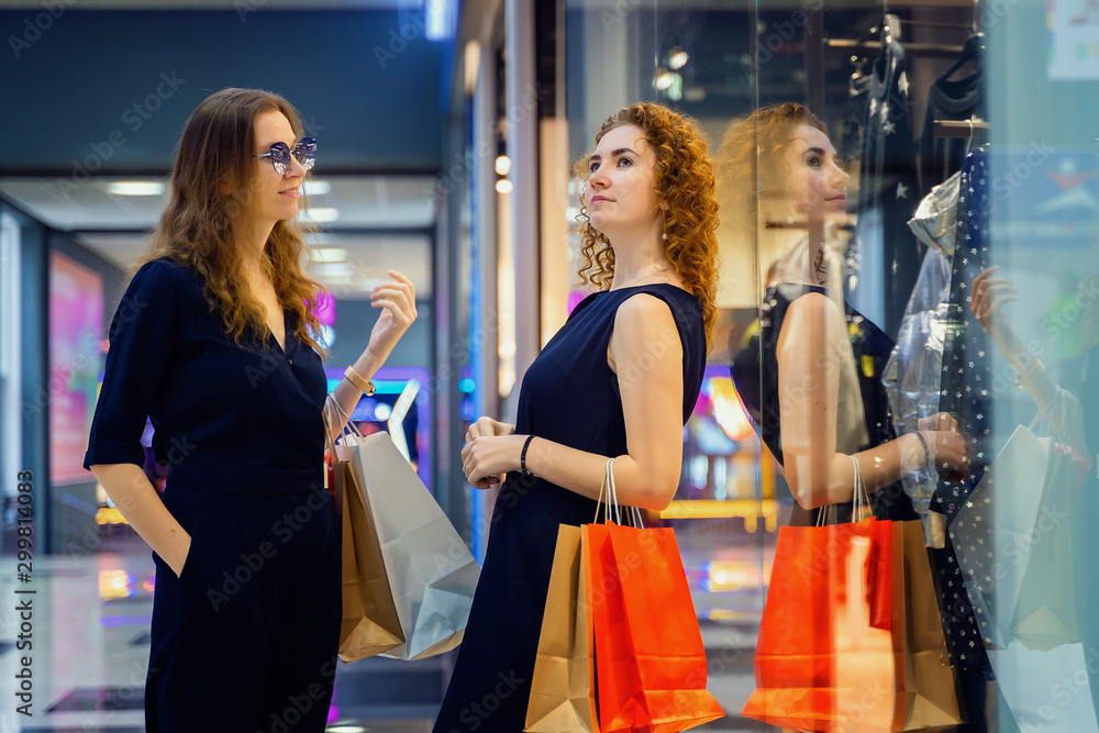 Two young beautiful girls are standing in a large shopping center near a shop window with packages and talking Two young beautiful girls are standing in a large shopping center near a shop window with packages and talking