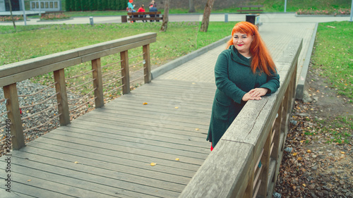 Shy old, red-haired woman leaning on handhold in park. Charming pretty woman thoughtfully looking at camera