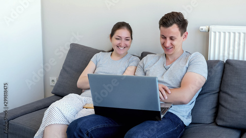 Young couple, man and woman are watching comedy movie on laptop and laughing sitting on sofa. They are in their modern apartment.