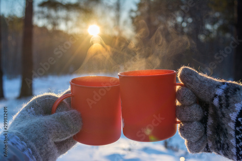 young man and young woman in knitted wool mittens with mugs of hot drink on sunny frosty day on forest background photo