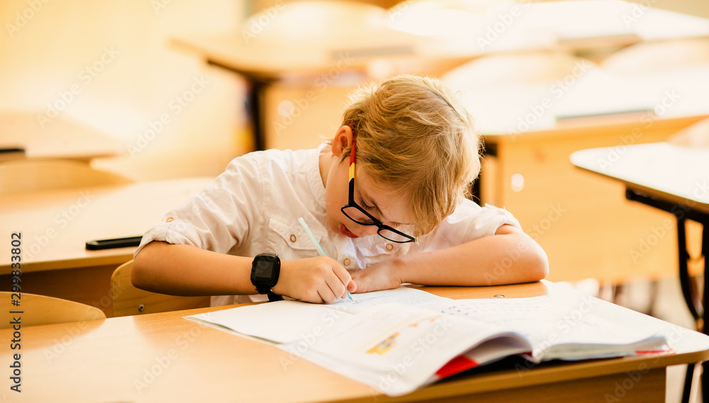 Seven years old child with glasses writing his homework at school. Boy studing at table on class background Seven years old child with glasses writing his homework at school. Boy studing at table on class background