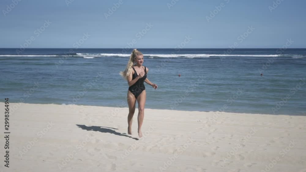 Young glamour woman with blond hair in small black shine dress and swimsuit running on white sand beach near ocean. With view of coastline and sky. Young glamour woman with blond hair in small black shine dress and swimsuit running on white sand beach near ocean. With view of coastline and sky.