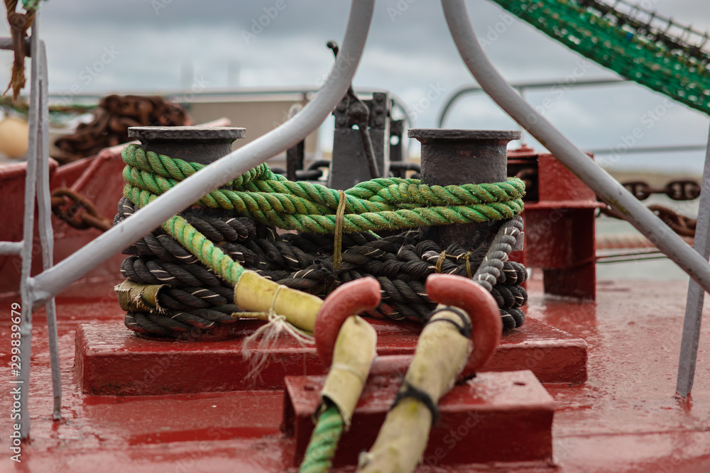 A closeup of a fishing ship. With green ropes, the ship is tied to a bollard. The ship's floor is painted red and the rail is gray.