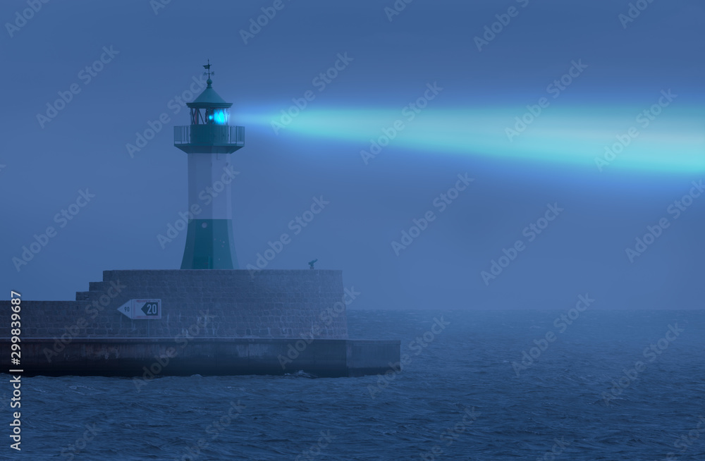 A small lighthouse with green light at the harbor entrance of the German Baltic Harbor Sassnitz on the island of Ruegen at night in fog. The light beam is visible in the fog.