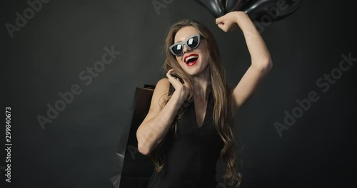 Pretty young woman in sunglasses and dress holding shopping bag and air balloons smiling to camera, waiting for black friday sales