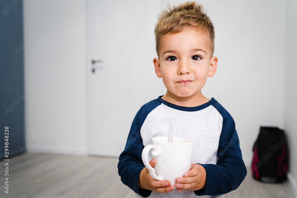 Small boy standing with a white cup of tea or milk holding in his hands at home Small boy standing with a white cup of tea or milk holding in his hands at home
