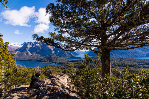 Young couple resting on the rocks beneath the shadow of a coihue tree against Andes range and Nahuel Huapi lake in Bariloche, Patagonia, Argentina 