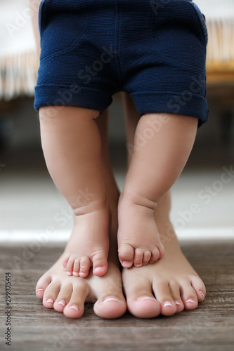 The legs of the baby stand on the feet of the mother, the mother supports the little son.Adorable baby's and mother's feet on floor at living room. Legs of mother and baby. First steps. Mom teaches he