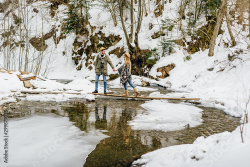 Couple of travelers overcome obstacle, mountain river in winter.