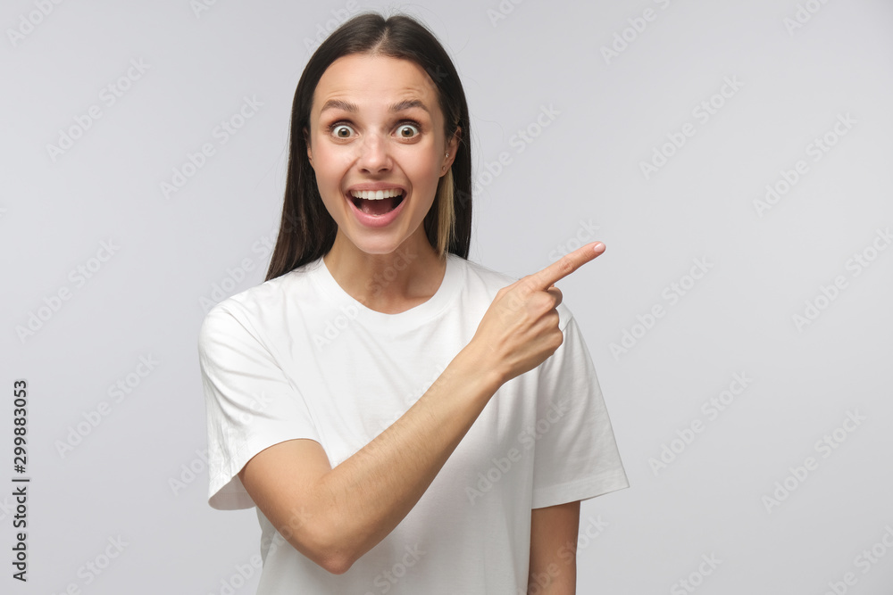 Young surprised woman in white t-shirt pointing right with her finger, shouting WOW, isolated on gray background Young surprised woman in white t-shirt pointing right with her finger, shouting WOW, isolated on gray background