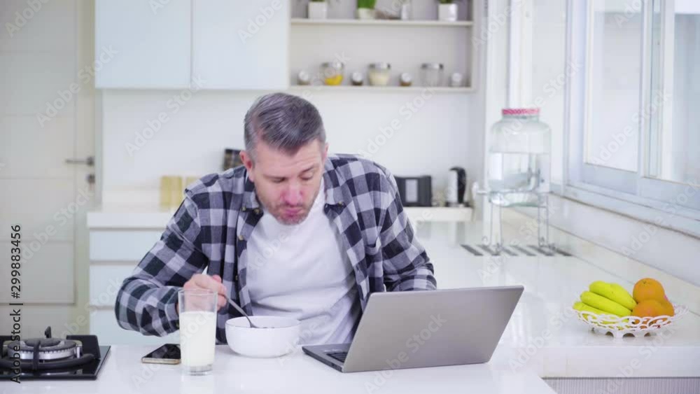 Attractive caucasian man working with laptop while breakfast with cereal in the kitchen at home. Shot in 4k resolution Attractive caucasian man working with laptop while breakfast with cereal in the kitchen at home. Shot in 4k resolution