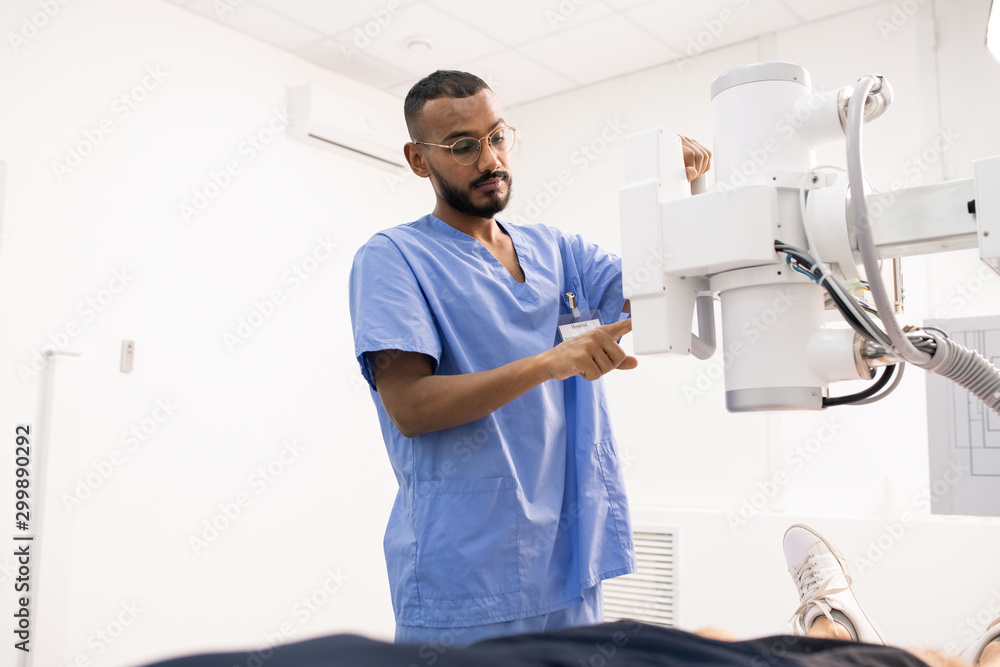 Bearded young mixed-race man in blue uniform using new medical equipment - obrazy, fototapety, plakaty 