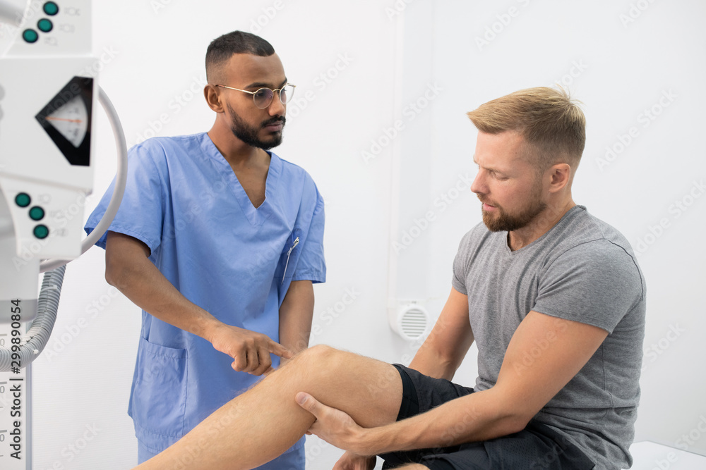 Young doctor in uniform consulting sportsman while pointing at his sick leg Young doctor in uniform consulting sportsman while pointing at his sick leg