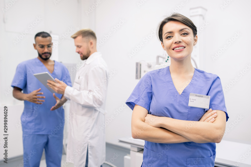 Happy young cross-armed clinician in uniform standing in front of camera Happy young cross-armed clinician in uniform standing in front of camera
