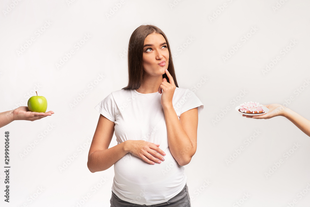 Puzzled Pregnant Girl Choosing Between Apple And Donut, White Background Puzzled Pregnant Girl Choosing Between Apple And Donut, White Background
