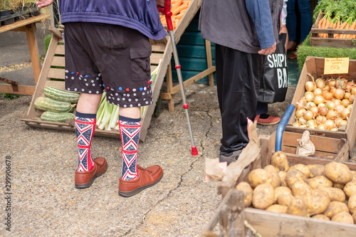 Waist down image of an endearing man seen wearing Union Jack socks while waiting to be served at a grocers. Various vegetables can be seen on display.